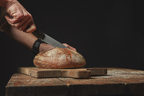 man slicing fresh organic bread on a wooden board, on an old brown table, on a dark background