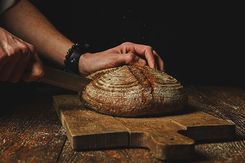 Hands of a young man cut fresh rye bread on a wooden cutting board, against a dark background. Against the background of a wooden old table