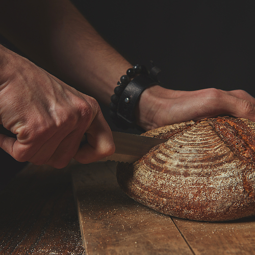 close-up on an old brown table, a man cuts a round dark bread on a wooden board against a dark background