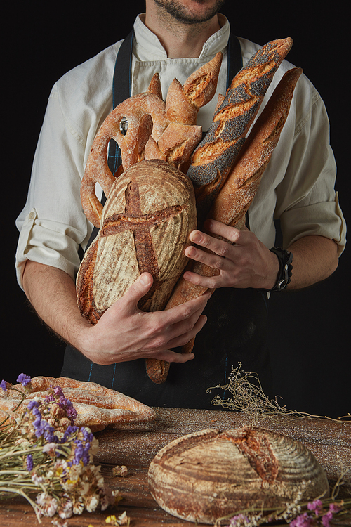 Hand of a baker holding fresh organic bread and baguette on a dark background wooden table with dry flowers and round bread