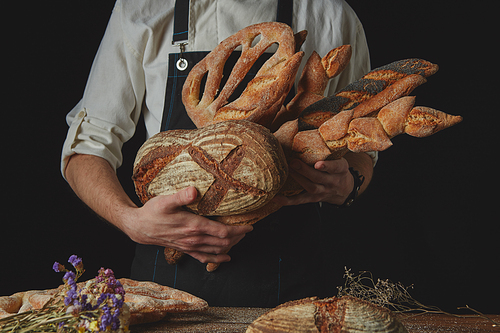 The baker's hands hold many different kinds of bread near a wooden table with dried flowers