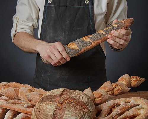 Baker holds a baguette with poppy seeds on a dark background and a variety of bread on the table