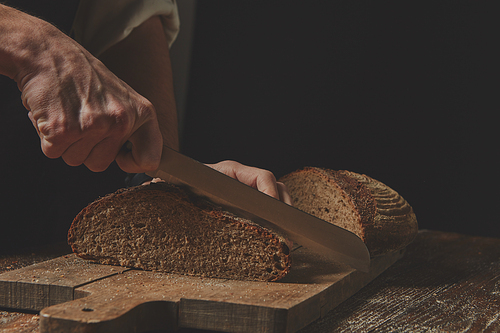 Hands of a baker cut fresh rye bread on a wooden background
