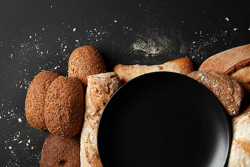 Top view of black plate represented among different kinds of breads over black background. Plate's copy space may be used for noting your ideas, emotions, feelings.