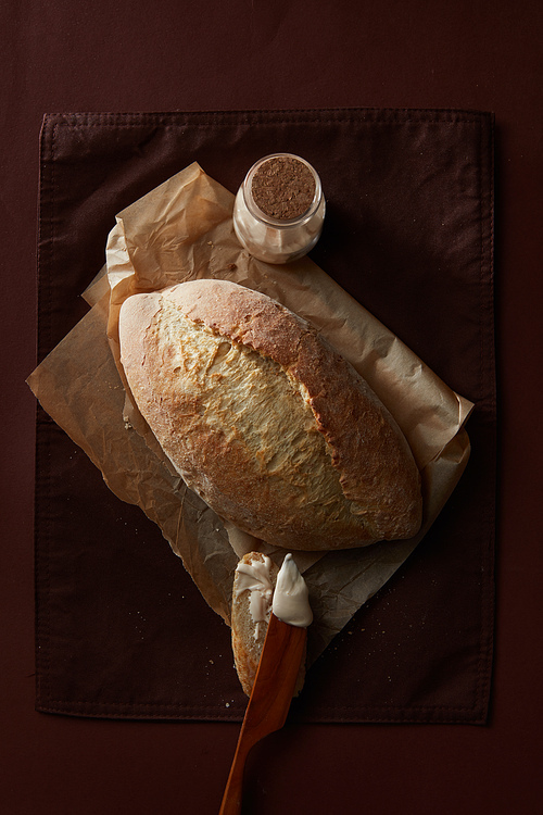 Fresh wheat bread on parchment, slice of bread with knife and butter and a jar with flour on a black background, flat lay