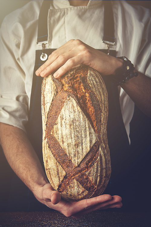 Men's hands on a black background hold a delicious fresh oval bread. Tonned photo