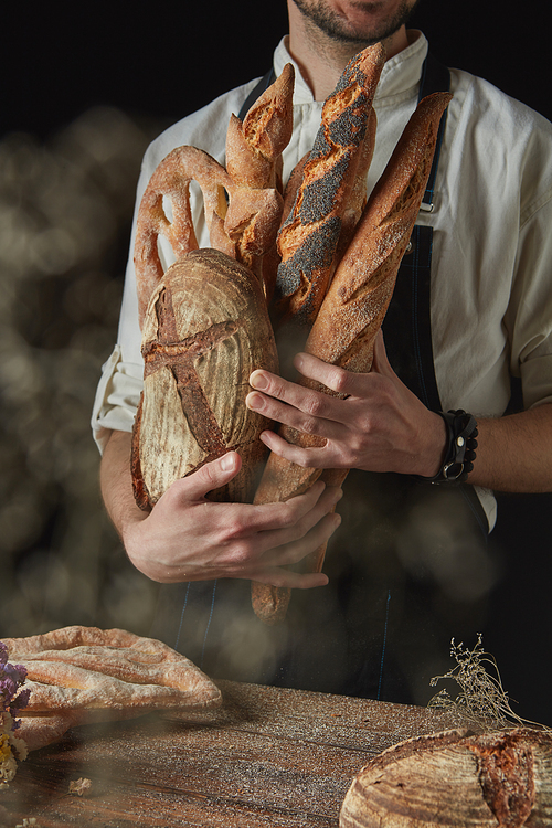 Fresh crunchy bread in the hands of a baker on a dark background near a wooden table with round bread