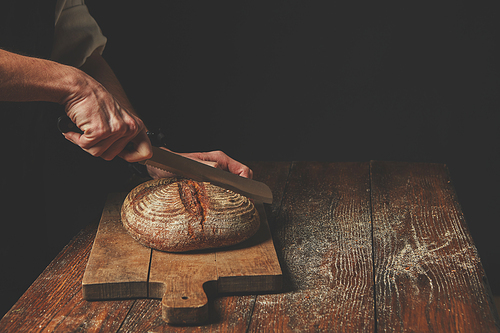 Baker's hands cut fresh round bread on a dark background