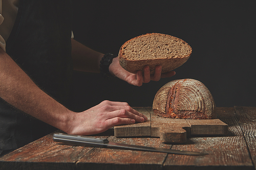 Baker keeps half freshly baked organic bread on a dark background
