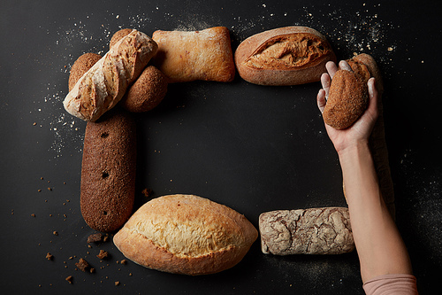 Top view of frame of different bread sorts over black background. Woman's hand holding bun on palm. Copy space in middle may be used for your ideas, emotions.
