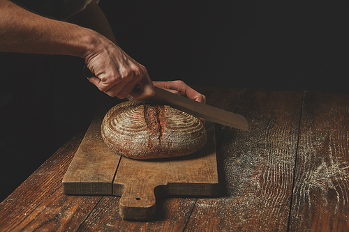 Men's hands cut round rye bread on a wooden brown cutting board