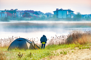 Green tent and a man near river with morning mist on the water. Outdoor travel landscape