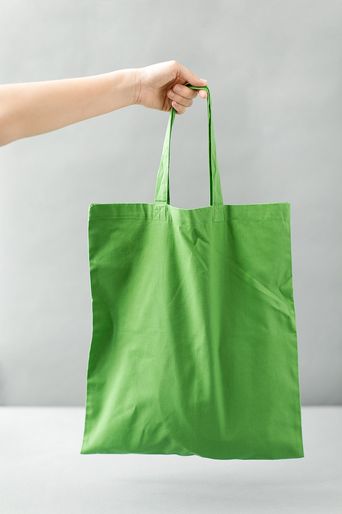 consumerism and eco friendly concept - hand holding reusable canvas bag for food shopping on grey background