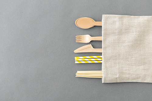 recycling and eco friendly concept - wooden disposable spoons, forks, knives, chopsticks and paper straws with canvas napkin on grey background