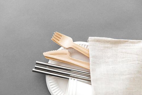 recycling and eco friendly concept - set of wooden forks, knives, paper cups and metallic straws with napkin on plate on grey background