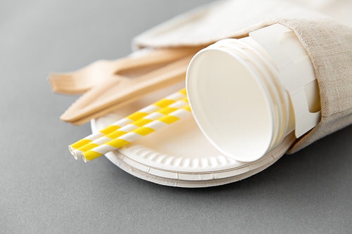 recycling and eco friendly concept - wooden disposable forks, knives, paper cups and straws with canvas napkin on grey background