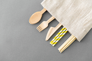 recycling and eco friendly concept - wooden disposable spoons, forks, knives, chopsticks and paper straws with canvas napkin on grey background