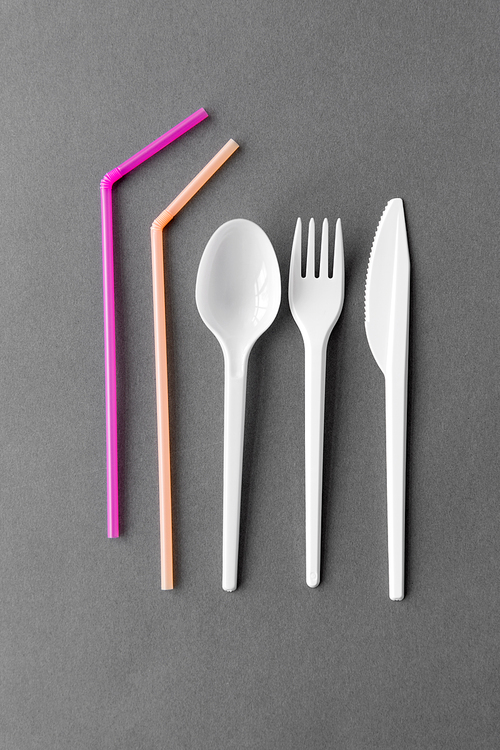 eating, recycling and ecology concept - white disposable plastic fork, knife and spoon with pink straws on grey background