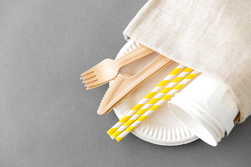 recycling and eco friendly concept - wooden disposable forks, knives, paper cups and straws with canvas napkin on grey background