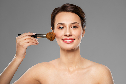 cosmetics and beauty concept - beautiful woman with makeup brush applying blusher over grey background