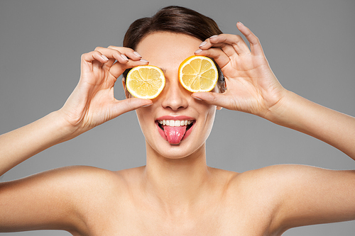beauty, skin care and detox concept - beautiful woman making eye mask of lemon slices and showing her tongue over grey background