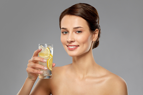 beauty and detox concept - woman drinking fresh water with lemon and ice over grey background