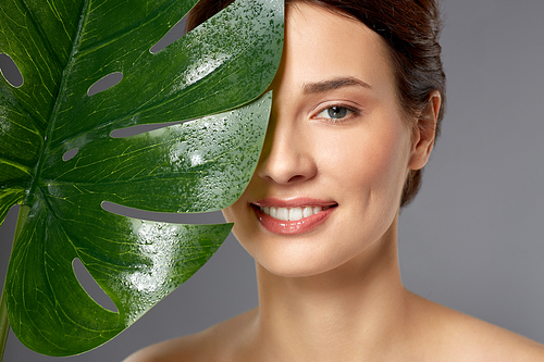 beauty, bodycare and people concept - beautiful young woman face with green monstera leaf over grey background