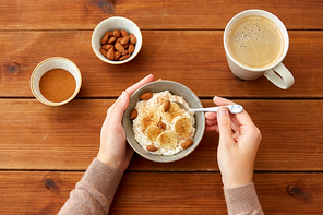 food and breakfast concept - hands of woman eating oatmeal porridge in bowl with sliced banana, almond nuts and cinnamon and cup of coffee on wooden table