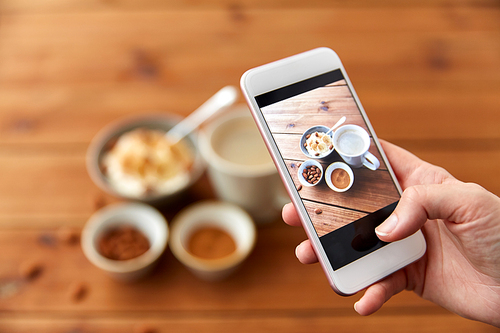 food, eating and breakfast concept - hand of woman taking picture of porridge in bowl, almond nuts, cinnamon and cup of coffee on wooden table with smartphone