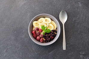 food and breakfast concept - oatmeal cereals in bowl with wild berries, banana and spoon on slate stone table