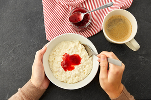 food and breakfast concept - hands of woman with spoon eating porridge in bowl with fruit jam and cup of coffee on slate stone table