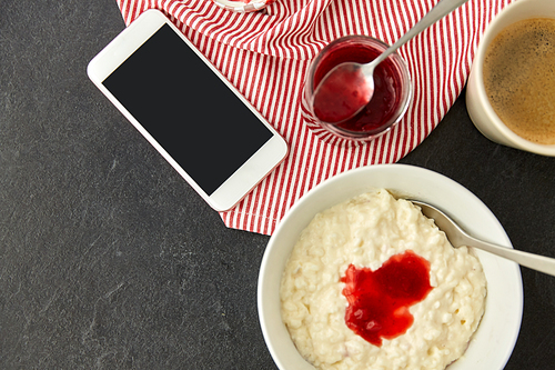 food and breakfast concept - porridge in bowl, smartphone, jam in mason jar, spoon and cup of coffee on slate stone table