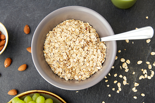 food, eating and breakfast concept - raw oatmeal cereals in bowl with spoon on slate stone table