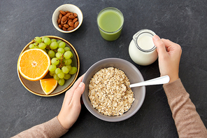 food and breakfast concept - hands of woman eating oatmeal cereals in bowl, fruits, almond nuts, glass of juice and jug with milk on slate stone table