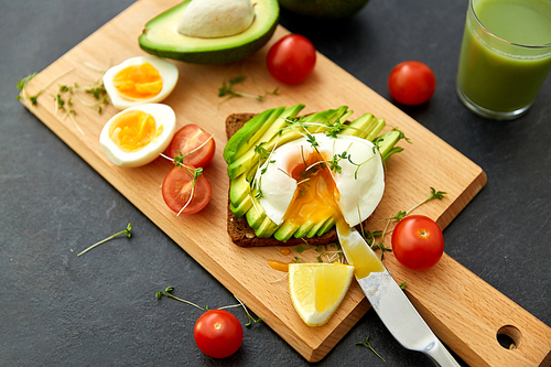 food, eating and breakfast concept - toast bread with sliced avocado, pouched egg, cherry tomatoes and table knife on wooden cutting board