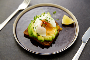 food, eating and breakfast concept - toast bread with sliced avocado, pouched egg and greens on ceramic plate with fork and knife