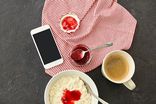 food and breakfast concept - porridge in bowl, smartphone, jam in mason jar, spoon and cup of coffee on slate stone table