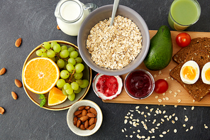 food, eating and breakfast concept - oatmeal cereals in bowl, fruits, almond nuts, glass of juice and jug with milk on slate stone table