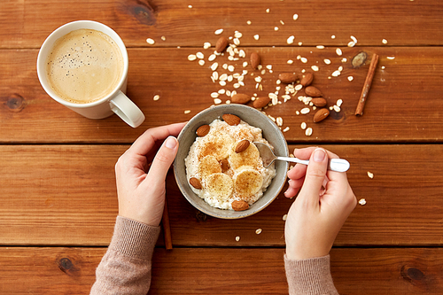 food and breakfast concept - hands of woman eating oatmeal porridge in bowl with sliced banana, almond nuts and cinnamon and cup of coffee on wooden table