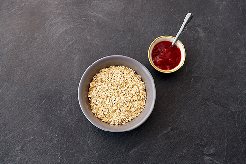 food and breakfast concept - oatmeal cereals in bowl and cup of fruit jam with spoon on slate stone table