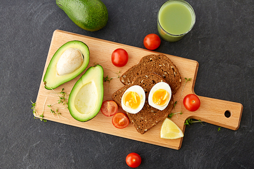 food, eating and breakfast concept - toast bread with eggs, cherry tomatoes, avocado and greens on wooden cutting board