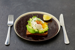 food, eating and breakfast concept - toast bread with sliced avocado, pouched egg and greens on ceramic plate with fork and knife