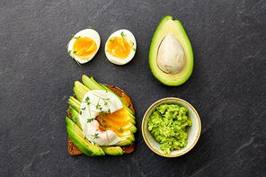 food, eating and breakfast concept - toast bread with sliced avocado, pouched egg and greens on slate stone table