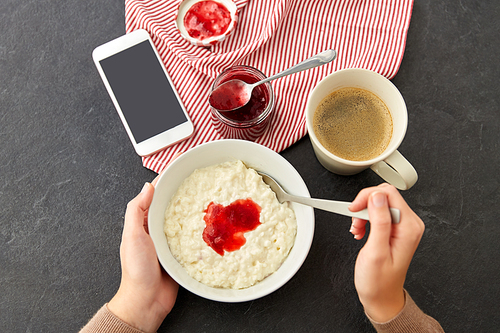 food and breakfast concept - hands of woman eating porridge in bowl, smartphone, jam in mason jar, spoon and cup of coffee on slate stone table