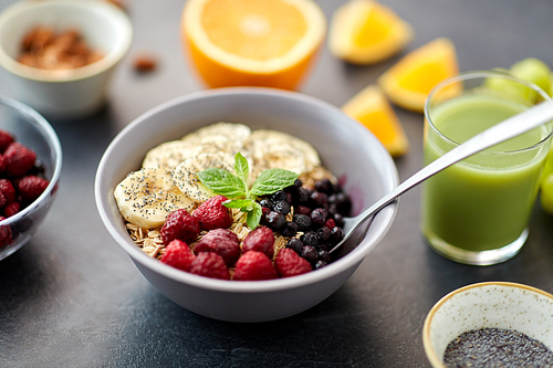 food and breakfast concept - oatmeal cereals in bowl with wild berries, banana, spoon and glass of juice on slate stone table