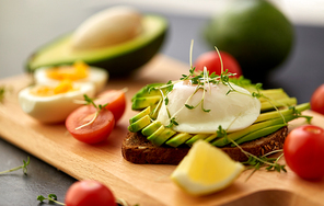 food, eating and breakfast concept - toast bread with sliced avocado, pouched eggs, cherry tomatoes and greens on wooden cutting board
