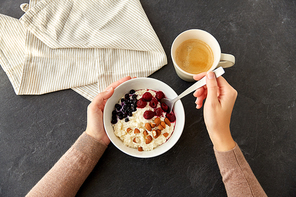 food and breakfast concept - hands of woman with spoon eating porridge in bowl with wild berries, almond nuts and cup of coffee on slate stone table
