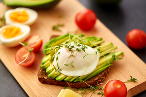 food, eating and breakfast concept - toast bread with sliced avocado, pouched egg, cherry tomatoes and greens on wooden cutting board