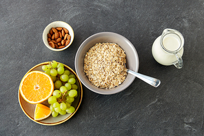 food and breakfast concept - oatmeal cereals in bowl, fruits, almond nuts and jug with milk on slate stone table