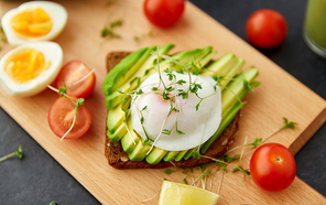 food, eating and breakfast concept - toast bread with sliced avocado, pouched egg, cherry tomatoes and greens on wooden cutting board